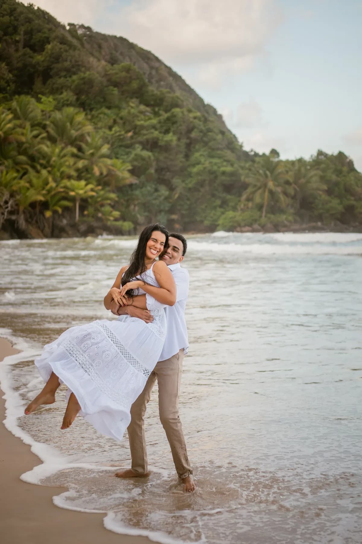 Isaque erguendo Beatriz no colo na beira do mar, com o morro verde ao fundo