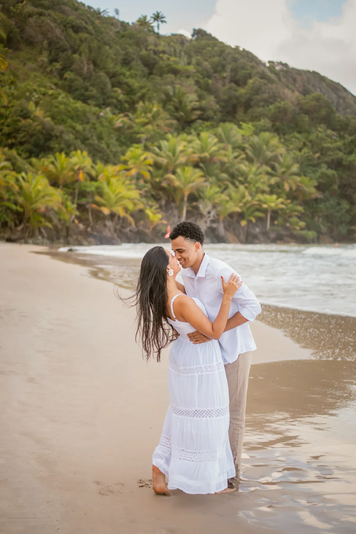 O casal abraçado na areia, prestes a se beijar, com os coqueiros ao fundo
