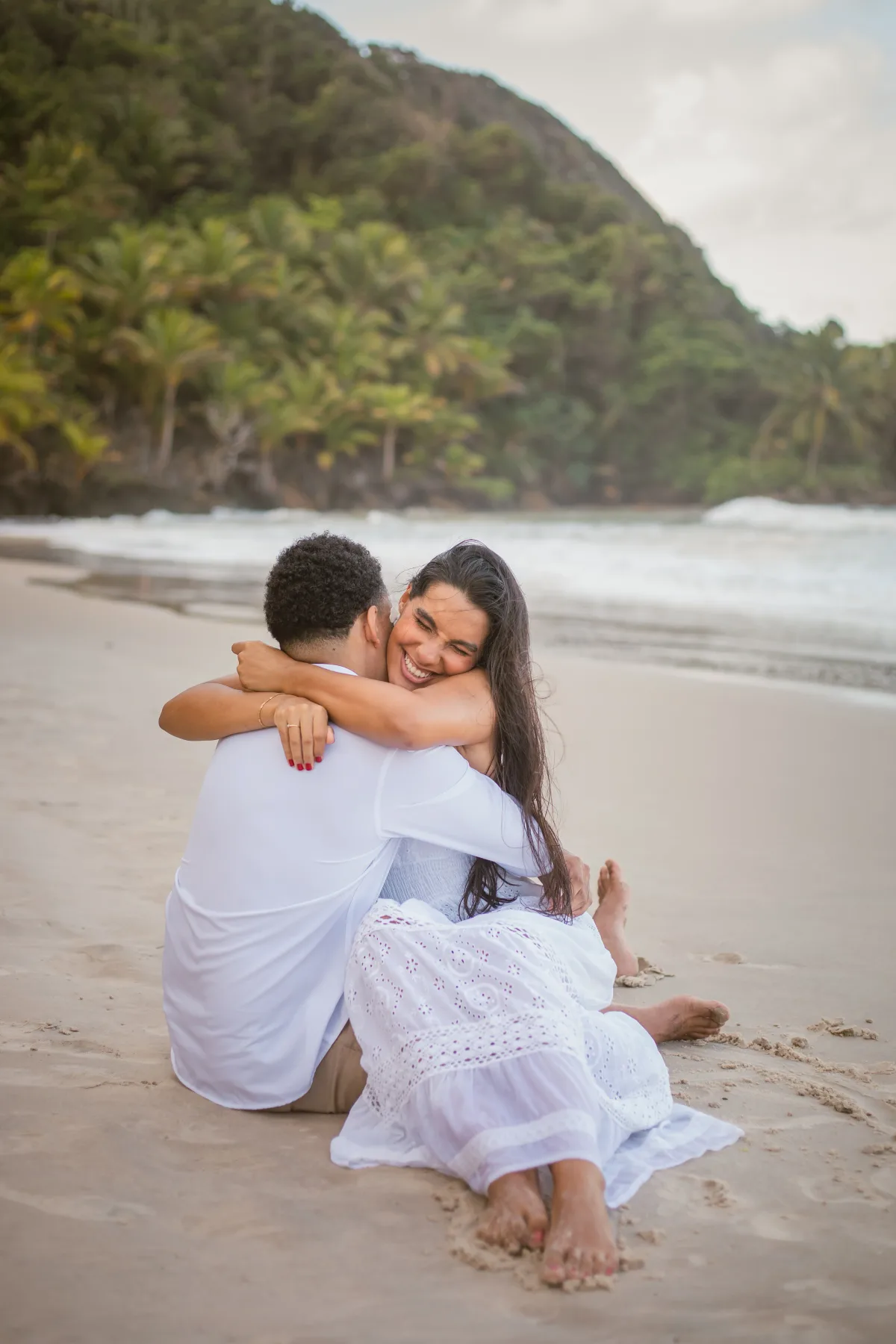 O casal sentado na areia, abraçado e rindo
