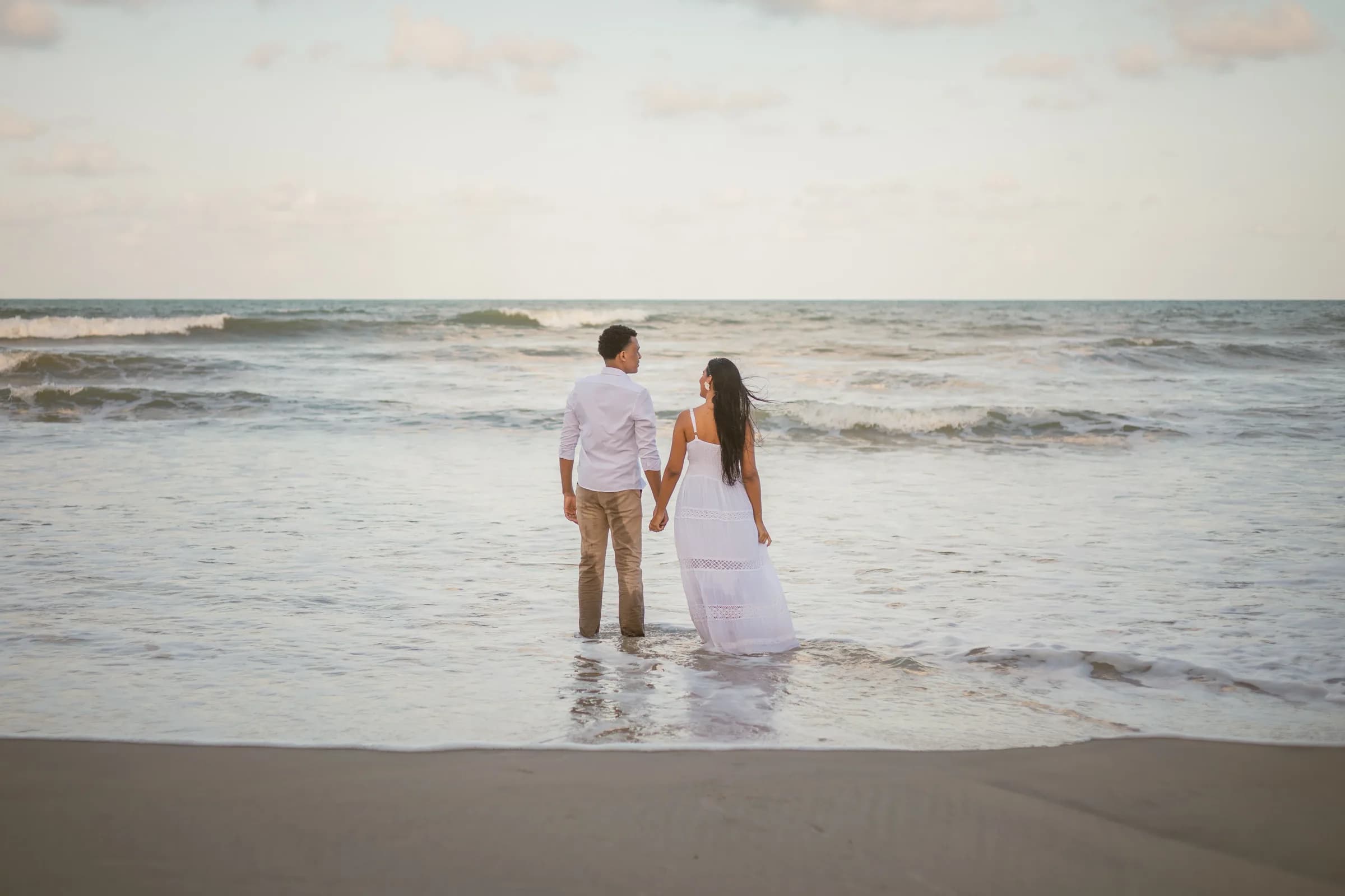 O casal de mãos dadas dentro do mar, de costas, olhando um para o outro