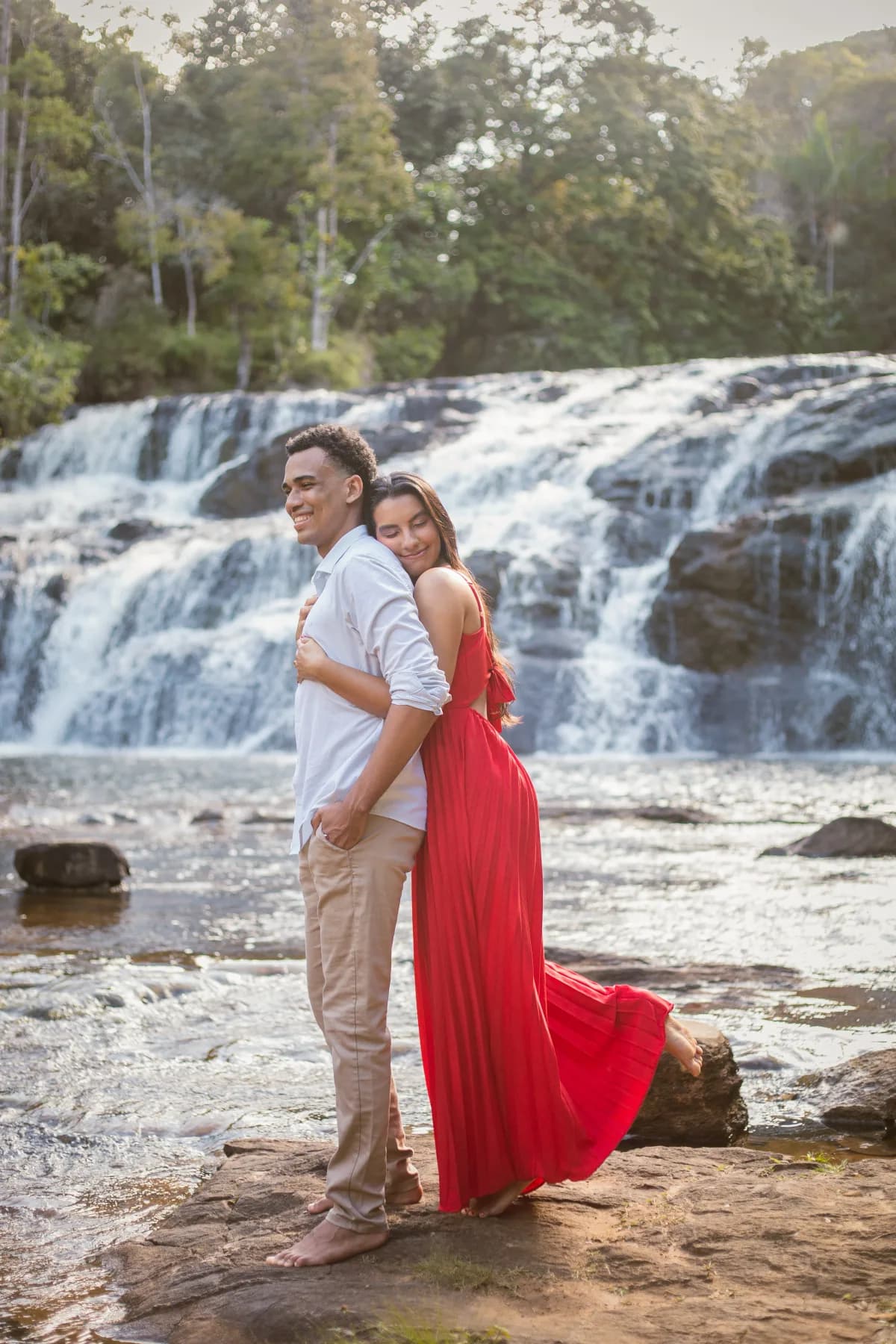O casal encostado de costas um no outro diante da cachoeira, ela de vestido vermelho