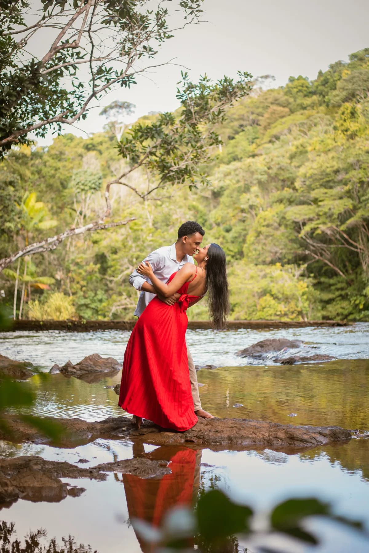 O casal se beijando sobre uma pedra à beira do rio, ela de vestido vermelho longo